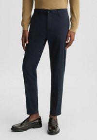 Marineblaue Hose mit schmaler Passform, aus einem glatten Stoff gefertigt. Mit Knopfverschluss und Fronttaschen, kombiniert mit dunkelbraunen Slippers.