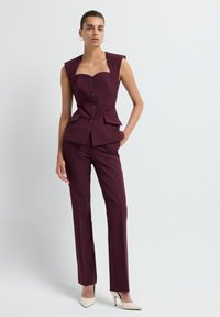 Costume ajusté bordeaux avec un décolleté carré, des détails de boutons et des poches à rabat. Pantalons taille haute, texture lisse et talons aiguilles blancs.