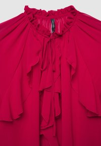 Blouse rouge avec un col à volants, en tissu doux, et fermeture par lien. De délicats volants descendent sur le devant, ajoutant texture et détail.