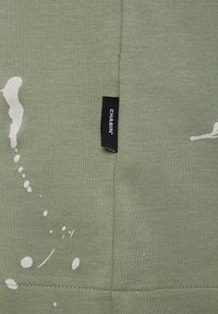 Green cotton fabric with white splatter design. Features a black branded label "CHASIN’" attached near the seam. Smooth texture, casual style.