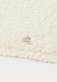 Tessuto di colore crema con texture, caratterizzato da un design shaggy, con un piccolo logo "CK" in argento ricamato sul bordo.