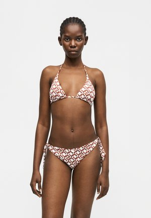 Donna con capelli intrecciati che indossa un bikini a fascia marrone con motivi bianchi e slip con laccetti laterali, in piedi davanti a uno sfondo chiaro e uniforme.