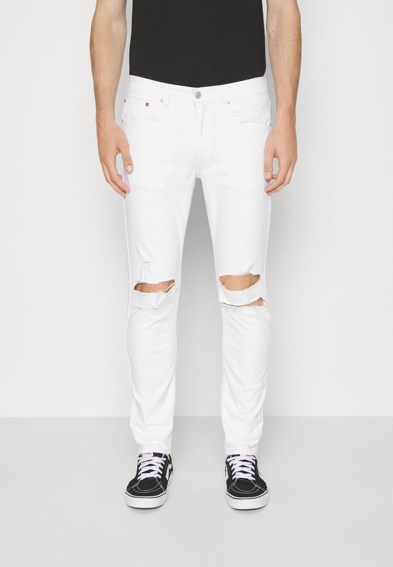Personne portant un jean slim blanc déchiré avec des trous aux genoux, une chemise noire et des baskets noires et blanches, debout contre un mur blanc.