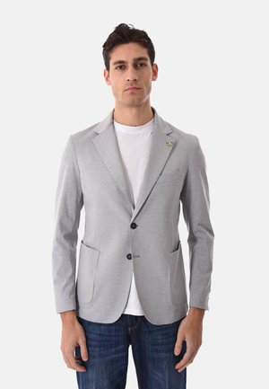 Uomo con blazer grigio chiaro sopra una t-shirt bianca e jeans blu, in piedi davanti a uno sfondo bianco uniforme.