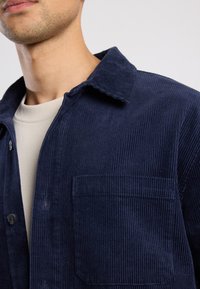 Navy corduroy shirt with a shirt collar, front pocket, and button closure. The fabric has a ribbed texture.