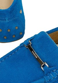 Niebieskie zamszowe mokasyny slip-on o teksturowanym wykończeniu, z metalicznym akcentem w postaci logo oraz okrągłymi ćwiekami na pięcie dla dodatkowego efektu.