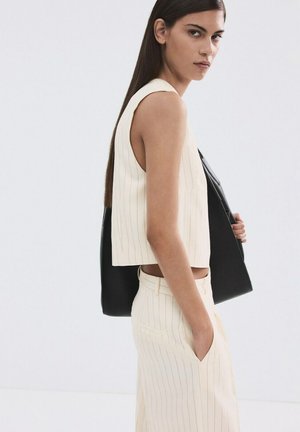 Donna con capelli lunghi e scuri che indossa un top senza maniche a righe color crema e pantaloni abbinati, con una giacca nera appoggiata sulla spalla.