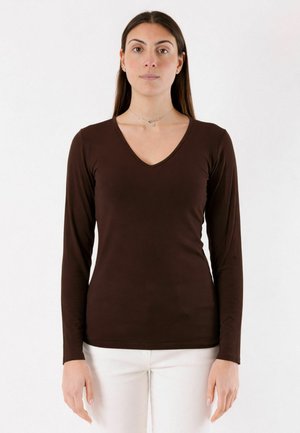 Donna che indossa una maglia a maniche lunghe a collo a V marrone scuro aderente e pantaloni bianchi, in piedi dritta su uno sfondo bianco uniforme.