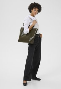 Sac fourre-tout de couleur vert olive, brillant, de forme rectangulaire avec des poignées courtes. Le mannequin porte une chemise blanche à col et un jean noir taille haute.