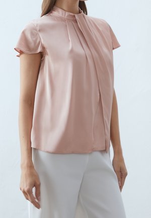 Woman wearing a blush pink satin short-sleeve blouse with pleated front detail and white high-waist trousers.