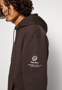 Brun hættebluse med en forkortet lomme, der har hvid tekst og grafik på ærmet. Stoffet ser blødt ud med en glat tekstur.