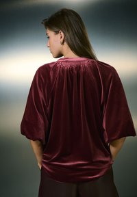 Haut en velours bordeaux avec un détail froncé à l'arrière, manches courtes et texture lisse. Conçu pour une coupe décontractée.