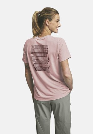 Femme souriante aux cheveux blonds attachés en queue de cheval, portant un t-shirt rose avec le texte vertical « Outdoor » dans le dos et un pantalon gris, les mains dans les poches.