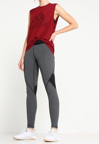 Punainen hihaton toppi, jossa on suuri musta Adidas-logo, yhdistettynä tiukkoihin harmaisiin legginsseihin, joissa on mustia yksityiskohtia ja geometrisia leikkauksia. Valkoiset urheilukengät.