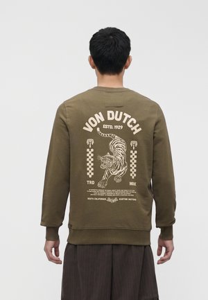 Mężczyzna w oliwkowej bluzie z nadrukiem w stylu vintage przedstawiającym tygrysa i tekstem "Von Dutch" na plecach, stojący na jednolitym jasnym tle.