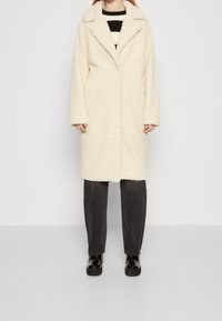 Abrigo texturizado color crema con cuello de muesca, mangas largas y cierre de un solo botón, usado sobre pantalones gris oscuro y zapatos negros.