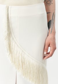 Jupe blanche avec un accent de frange diagonale en crème. Le tissu est lisse avec un design structuré, mettant en valeur le détail de la frange.