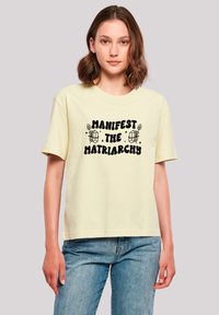Light yellow cotton t-shirt with short sleeves, featuring black text that reads "MANIFEST THE MATRIARCHY," surrounded by simple line drawings.