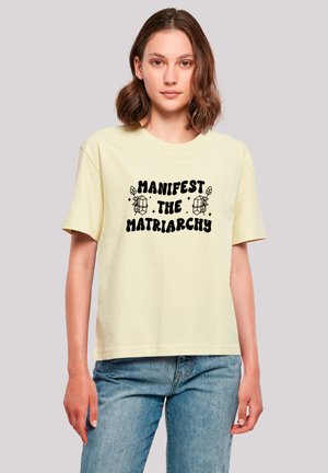 Light yellow cotton t-shirt with short sleeves, featuring black text that reads "MANIFEST THE MATRIARCHY," surrounded by simple line drawings.