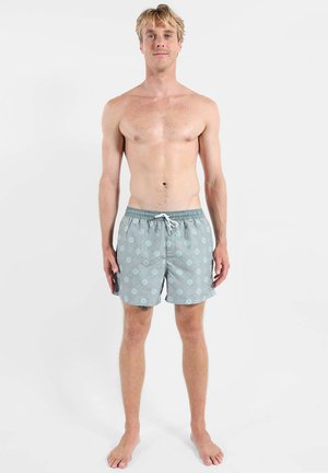 Jeune homme debout pieds nus portant un short de bain bleu clair à motifs avec un cordon blanc sur fond blanc.