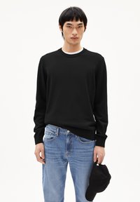 Schwarzer Pullover mit langen Ärmeln und Rundhalsausschnitt, kombiniert mit hellblauen Jeans, hält eine schwarze Baseballmütze. Weicher Stoff, lässige Passform.