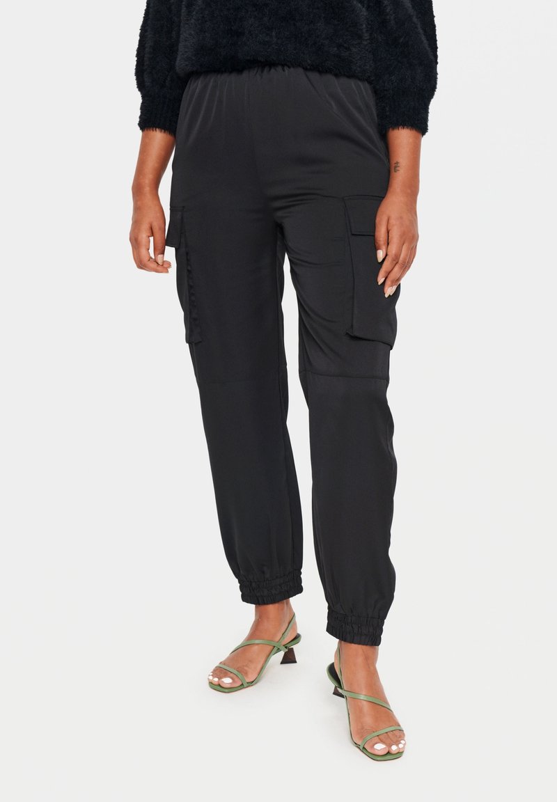 Femme portant un pantalon cargo noir avec des poignets élastiques et des sandales à talons vertes à fines lanières, debout devant un fond blanc.