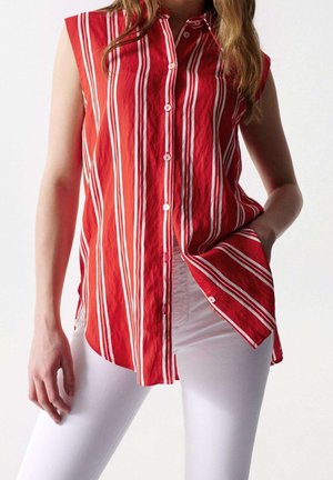 Chemise rouge sans manches à boutons avec des rayures verticales blanches portée avec un pantalon blanc, une main dans la poche, sur un fond uni.