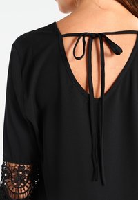 Svart topp med V-urringning bak som har knytband, korta ärmar prydda med intrikat svart spets och en slät tygkänsla.