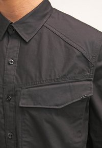 Camicia nera con bottoni realizzata in tessuto morbido, con colletto a punta, dettagli sulle spalle e taschino sul petto con patta.