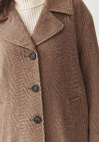 Ruskea herringbone-takki, jossa on lovellinen kaulus, neljä mustaa nappia ja sivutaskut. Valmistettu teksturoidusta kankaasta.