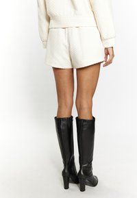 Tejbir colorú, texturált pamut sort, amelyet magas fekete bőr térdcsizmával párosítanak, blokk sarkú és cipzár részlettel.
