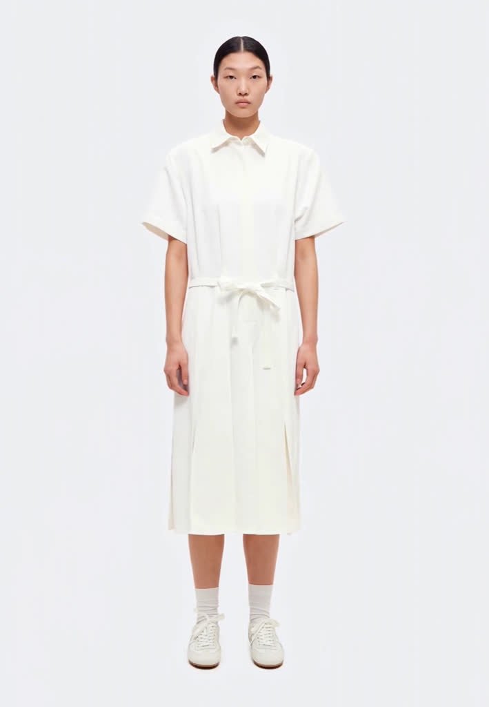 Femme aux cheveux foncés attachés en arrière, portant une robe chemise blanche à manches courtes avec une ceinture nouée à la taille et des baskets blanches, debout devant un fond blanc.