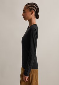 Profiel van een persoon met gevlochten haar, gekleed in een zwarte lange mouwen shirt en bruine broek, tegen een effen achtergrond.