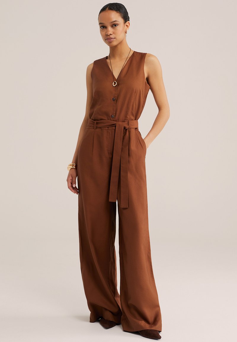 Jeune femme portant une combinaison marron sans manches à jambes larges, boutonnée devant, avec une ceinture à la taille, accessoirisée de bijoux en or, debout devant un fond uni.