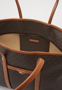 Sac cabas marron Michael Kors avec garniture et poignées en cuir camel, montrant une poche intérieure zippée et un détail de logo en or sur la poche extérieure.