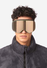 Skibrille mit übergroßem, verspiegeltem bronzefarbenem Glas und grünem Rahmen, getragen von einer Person in einer hochkragen, dunkelgrauen Jacke.