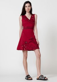 Rotes ärmelloses Kleid mit V-Ausschnitt, schwarzer floralem Stickerei und gemustertem Besatz an der Taille; kombiniert mit schwarzen verzierten Sandalen.
