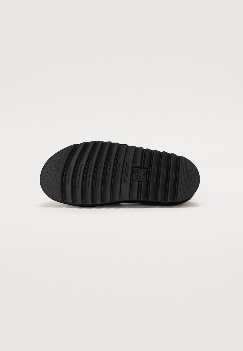 Black rubber shoe sole with horizontal and vertical grooves for traction. Central logo feature with a textured pattern. Rounded toe design.