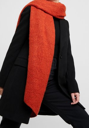 Personne vêtue entièrement de noir portant une écharpe épaisse et texturée de couleur orange vif enroulée autour du cou, sur fond gris uni.