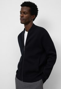 Svart blixtlåst bomberjacka med räfflad krage, sidofickor och en slät textur. Bärs över en vit skjorta, mot en neutral bakgrund.