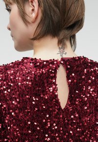 Κόκκινο φόρεμα με παγιέτες και λεπτομέρεια κλειδώματος στην πλάτη. Μαλακή υφή με γυαλιστερές κυκλικές παγιέτες που καλύπτουν το ύφασμα, αναδεικνύοντας μια εφαρμοστή σιλουέτα.