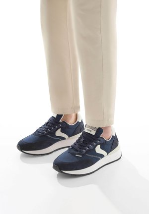 Navy Sneakers aus Mesh- und Wildledermaterial, weiße Akzente, gepolsterte Sohle und dunkle Schnürsenkel. Kombiniert mit hellbeigen Hosen.