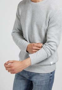 Pull en tricot gris avec un motif texturé, des poignets rayés et un accent triangulaire sur l'ourlet. Porté avec un jean bleu.