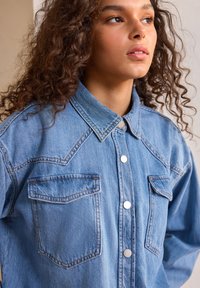Camicia in denim azzurro chiaro con chiusura a bottoni, colletto a punta, due tasche sul petto e dettagli di cucitura a contrasto.