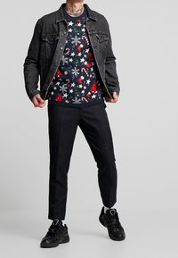 Svart denimjacka över en marinblå jultröja med godisstänger, träd och stjärnor. Ett par mörka byxor och svarta sneakers.