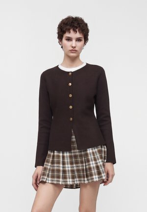 Jeune personne aux cheveux courts et bouclés porte un cardigan marron foncé boutonné et une jupe plissée à carreaux marron, blanc et noir.