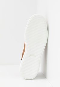 Semelle d'une chaussure marron avec une semelle extérieure en caoutchouc blanche rainurée, inclinée contre un fond blanc à angles.