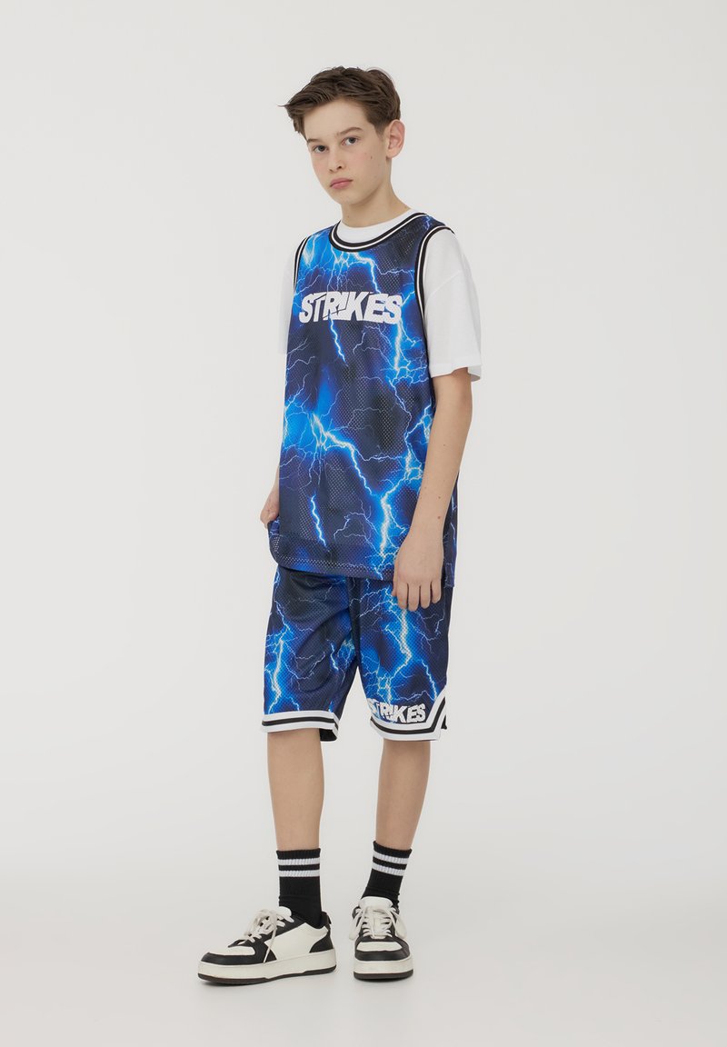 Maglia da basket blu con motivo a fulmine, abbinata a pantaloni corti. Accenti bianchi e bordi neri. Il modello indossa sneakers bianche e nere con calze a righe.