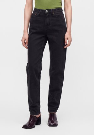 Person iført højtaljede sorte jeans kombineret med mørkerøde hælede sko og en olivengrøn kortærmet top, stående mod en ensfarvet baggrund.