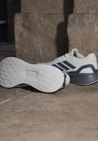 Weiße und marineblaue Sportschuhe mit strukturierter Sohle, die auf einem Steinboden vor einer Steinwand liegen.